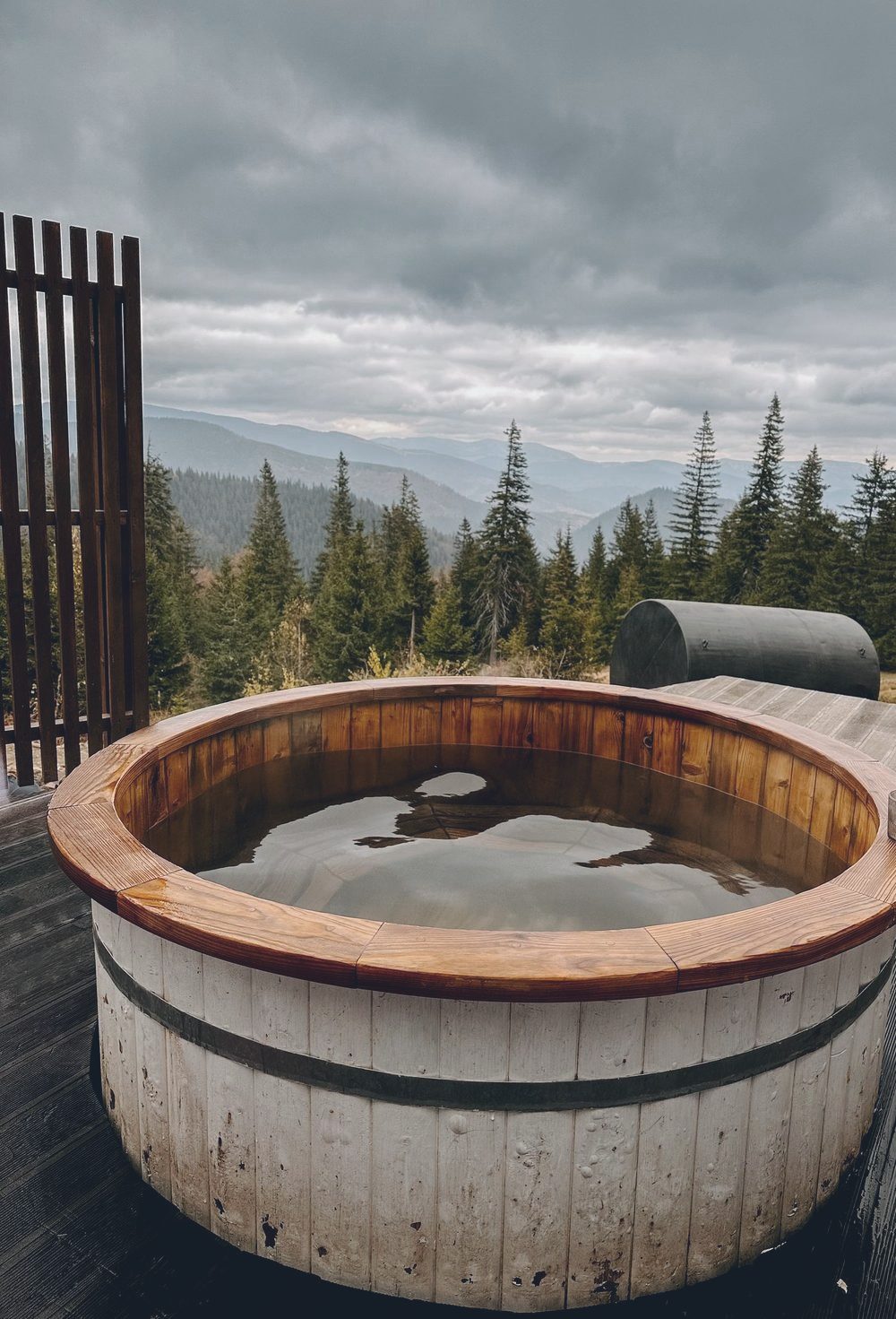 Houten hottub op een terras met uitzicht over naaldbos en heuvels, een ton-sauna erachter